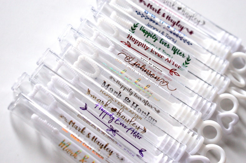 May include: Clear plastic bubble wands with white handles. Each wand has personalized text in various colors, including "Mark & Hayley" and "Happily Ever After." Ideal for weddings or celebrations.