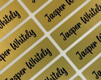 May include: A sheet of rectangular gold-colored adhesive labels arranged in a grid pattern. Each label features the name Jasper Whitely printed in a black, cursive script font. The labels have a metallic finish and rounded corners.