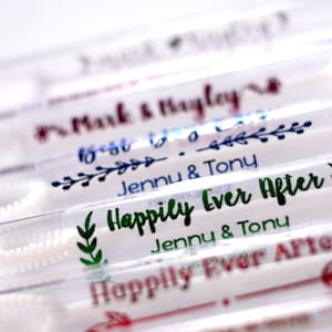 May include: Close-up of several clear bubble wands with personalized labels. The labels feature names and phrases like "Happily Ever After" and "Best Day Ever" in various colors, including green, blue, and red. The wands are arranged in a row.