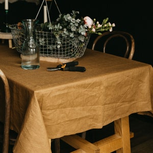 May include: A brown linen tablecloth covers a wooden table with a wire basket filled with greenery and pink flowers. A clear glass bottle and a pair of pruning shears are on the table.