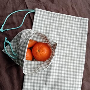 Op de afbeelding: Een herbruikbare grijze en witte gingham-tas voor producten met een turquoise trekkoord. De tas is gevuld met vier hele sinaasappelen.