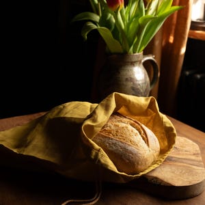 May include: A loaf of freshly baked bread is nestled inside a yellow linen bread bag on a wooden chopping board. A bouquet of red tulips in a ceramic vase is in the background.