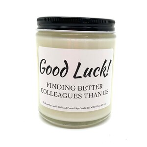 May include: A white soy candle in a glass jar with a black lid. The label reads "Good Luck! Finding Better Colleagues Than Us".