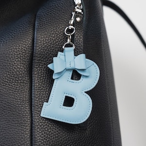 Può includere: Uno zaino in pelle nera con un portachiavi in pelle azzurra a forma di lettera "B". Il portachiavi ha un dettaglio a fiocco nella parte superiore ed è attaccato alla cerniera con una chiusura color argento. Lo zaino ha una chiusura con fibbia e spallacci regolabili.