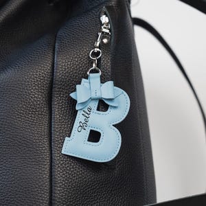 Peut inclure: Un sac à dos en cuir noir avec un porte-clés en forme de lettre "B" en cuir bleu clair. Le porte-clés a un nœud et le nom "Bella" en écriture cursive. Le sac a une fermeture éclair et une boucle argentées.