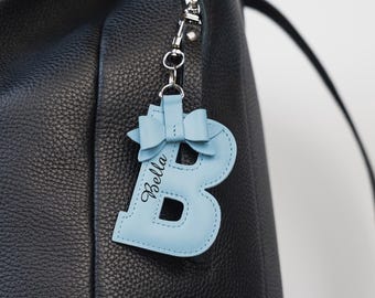 Peut inclure: Un sac à dos en cuir noir avec un porte-clés en forme de lettre "B" en cuir bleu clair. Le porte-clés a un nœud et le nom "Bella" en écriture cursive. Le sac a une fermeture éclair et une boucle argentées.