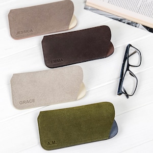 Puede incluir: Cuatro estuches de gafas de ante en tonos marrón, beige y verde oliva, cada uno con un interior en contraste. Los estuches están personalizados con nombres o iniciales: "JESSICA", "EMMA", "GRACE" y "A.M.". Un par de gafas con montura negra descansa cerca.