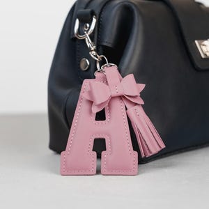 May include: A pink leather letter "A" keychain with a bow and tassel, attached to a black handbag. The keychain is a light pink color and has a stitched border. The handbag has a top handle and a silver clasp.