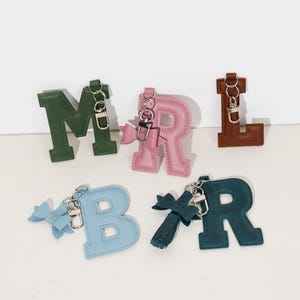 Può includere: Portachiavi a forma di lettera in vari colori, tra cui verde, rosa, marrone, blu e verde acqua. Ogni portachiavi presenta un fiocco o nappa e un fermaglio in metallo. Le lettere sono "M", "R", "L", "B" e "R".