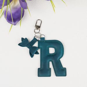 Könnte beinhalten: Türkisfarbener Leder-Schlüsselanhänger mit dem Buchstaben "R" und einer kleinen Schleife. Der Schlüsselanhänger hat einen silberfarbenen Verschluss und Schlüsselring. Der Hintergrund ist weiß mit lila Blumen.