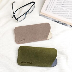 Peut inclure: Deux &eacute;tuis &agrave; lunettes personnalis&eacute;s, un taupe et un vert olive, avec les noms "JULES" et "KIMBERLY" estamp&eacute;s. Des lunettes &agrave; monture noire et un magazine ouvert sont &eacute;galement visibles sur une surface blanche textur&eacute;e.