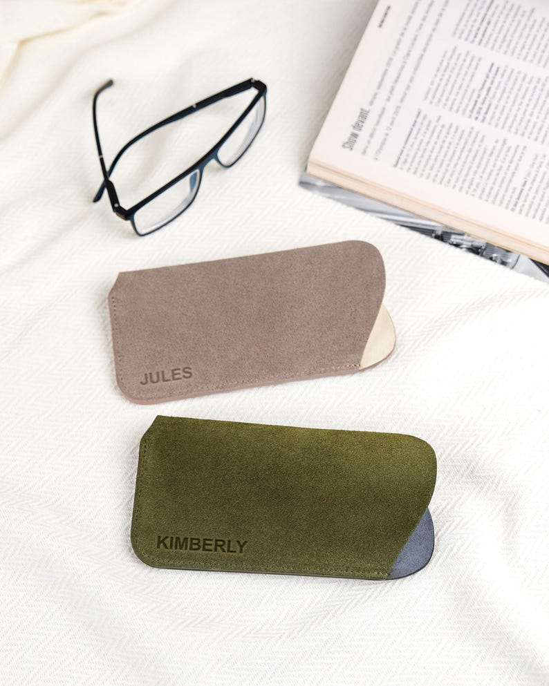 Peut inclure: Deux &eacute;tuis &agrave; lunettes personnalis&eacute;s, un taupe et un vert olive, avec les noms "JULES" et "KIMBERLY" estamp&eacute;s. Des lunettes &agrave; monture noire et un magazine ouvert sont &eacute;galement visibles sur une surface blanche textur&eacute;e.
