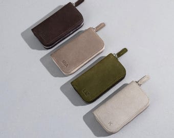 May include: Four suede key wallets in brown, tan, olive green, and cream. Each wallet has a zipper closure and a small loop. The wallets are embossed with initials: R.B., MIA, LIZ, and K.