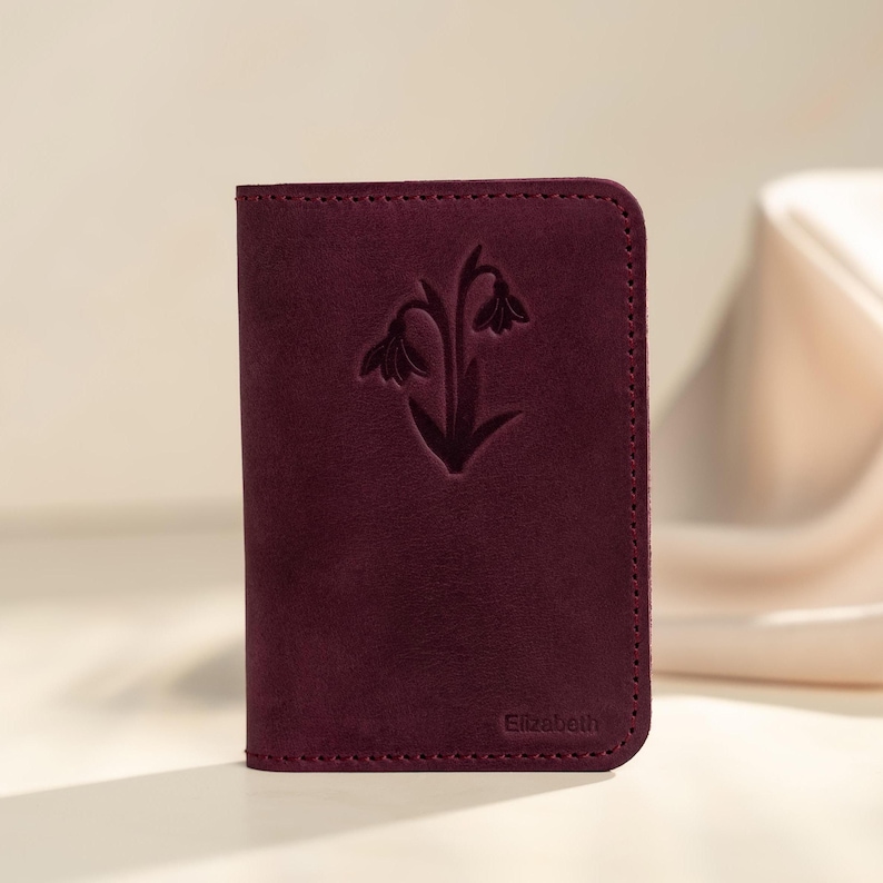 May include: A burgundy leather wallet with a floral design embossed on the front. The wallet has a stitched border and the name "Elizabeth" is embossed at the bottom. The wallet is rectangular and appears to be closed.
