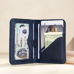 May include: Open navy blue leather wallet with multiple card slots and a clear ID window. The wallet contains a driver's license, credit cards, and a small amount of U.S. currency. The wallet is open, displaying its contents.
