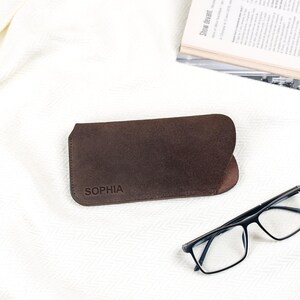 Peut inclure: Un &eacute;tui &agrave; lunettes en cuir marron avec le nom "SOPHIA" estamp&eacute; sur le devant. L'&eacute;tui est ouvert, r&eacute;v&eacute;lant un int&eacute;rieur sombre. Des lunettes &agrave; monture noire sont pos&eacute;es &agrave; c&ocirc;t&eacute; de l'&eacute;tui sur une surface blanche. Un magazine est en arri&egrave;re-plan.