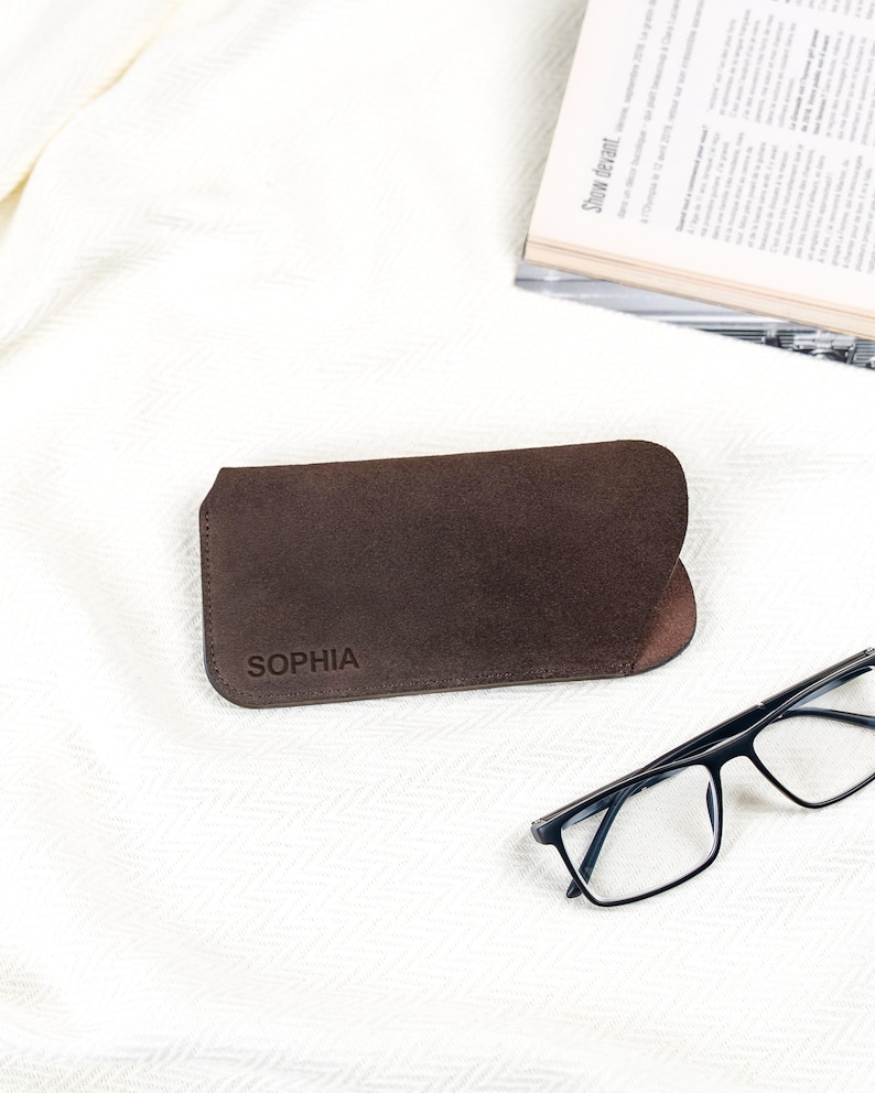 Peut inclure: Un &eacute;tui &agrave; lunettes en cuir marron avec le nom "SOPHIA" estamp&eacute; sur le devant. L'&eacute;tui est ouvert, r&eacute;v&eacute;lant un int&eacute;rieur sombre. Des lunettes &agrave; monture noire sont pos&eacute;es &agrave; c&ocirc;t&eacute; de l'&eacute;tui sur une surface blanche. Un magazine est en arri&egrave;re-plan.