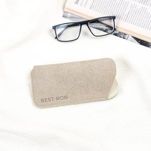 Peut inclure: Un &eacute;tui &agrave; lunettes beige avec le texte "BEST MOM" imprim&eacute; dessus. L'&eacute;tui est fait d'un mat&eacute;riau doux et est ouvert. Des lunettes &agrave; monture noire et un journal sont en arri&egrave;re-plan.