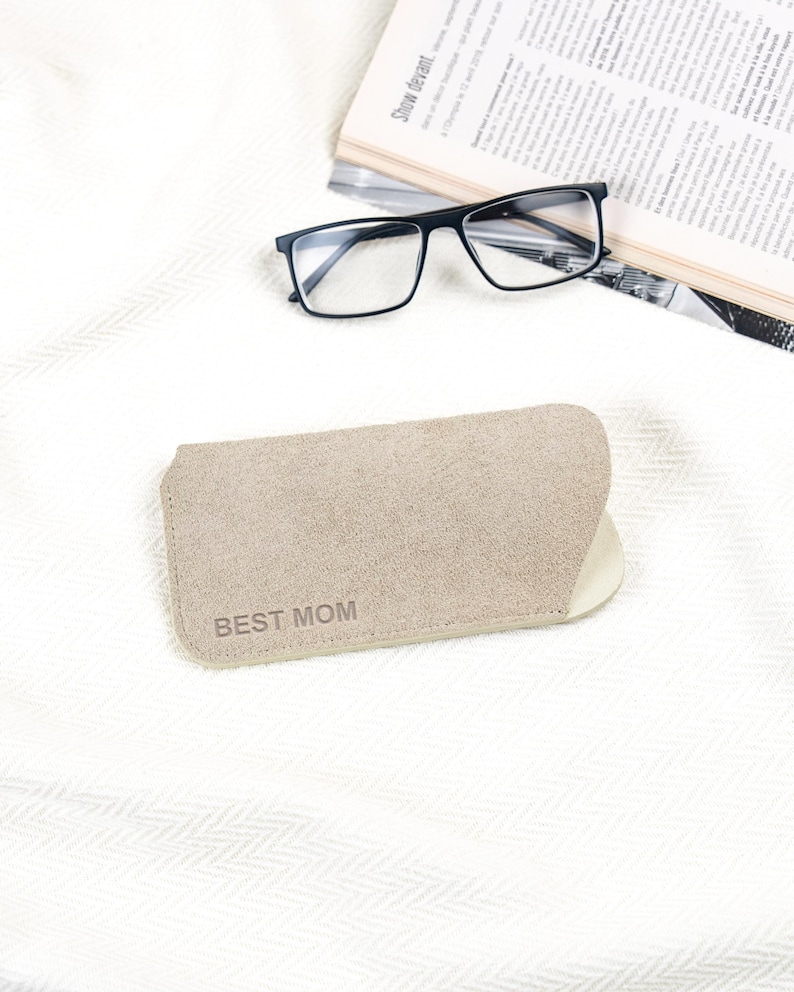 Peut inclure: Un &eacute;tui &agrave; lunettes beige avec le texte "BEST MOM" imprim&eacute; dessus. L'&eacute;tui est fait d'un mat&eacute;riau doux et est ouvert. Des lunettes &agrave; monture noire et un journal sont en arri&egrave;re-plan.