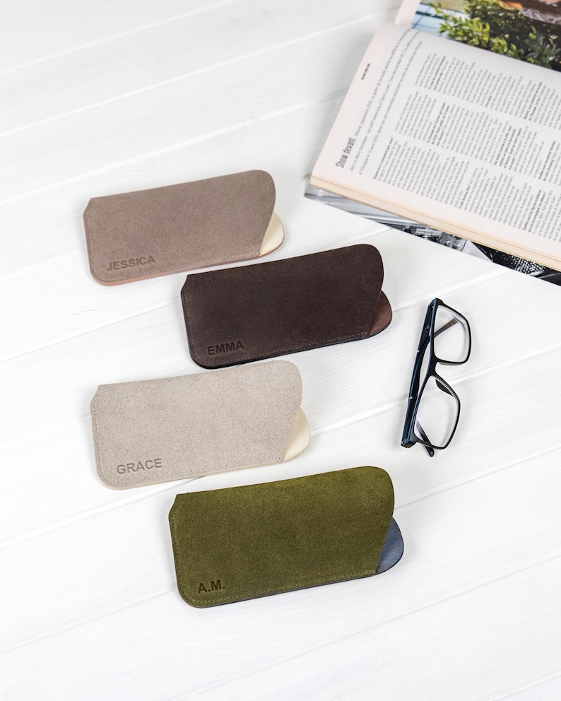 Peut inclure: Quatre &eacute;tuis &agrave; lunettes en daim dans des tons de marron, beige et vert olive, chacun avec un int&eacute;rieur contrast&eacute;. Les &eacute;tuis sont personnalis&eacute;s avec des noms ou des initiales : "JESSICA", "EMMA", "GRACE" et "A.M.". Une paire de lunettes &agrave; monture noire est pos&eacute;e &agrave; c&ocirc;t&eacute;.