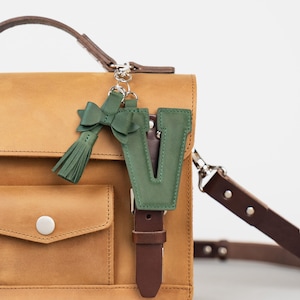 May include: A tan leather satchel bag with a front pocket and silver hardware. A green leather letter "V" keyring and a green bow with a tassel are attached to the bag's strap, adding a pop of colour to the design.