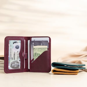 May include: Open burgundy leather wallet with multiple card slots and a clear ID window. The wallet is open, revealing cards and cash. Two additional wallets in teal and tan are stacked on the right.