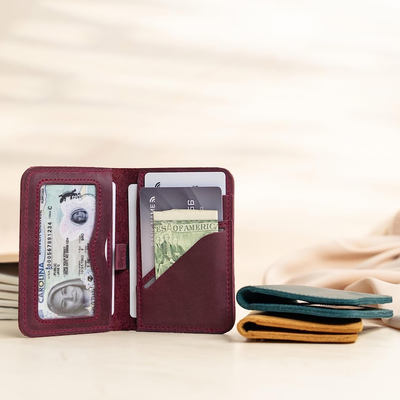May include: Open burgundy leather wallet with multiple card slots and a clear ID window. The wallet is open, revealing cards and cash. Two additional wallets in teal and tan are stacked on the right.
