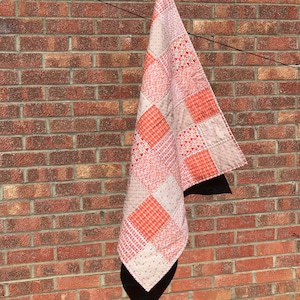 May include: A patchwork quilt with a variety of orange and white patterns. The quilt is hanging on a brick wall.