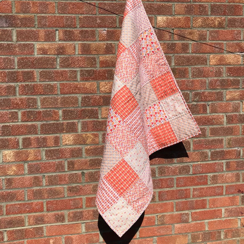 May include: A patchwork quilt with a variety of orange and white patterns. The quilt is hanging on a brick wall.