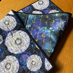 Op de afbeelding: Twee gestikte placemats met een hemels thema. De ene placemat heeft een donkerblauwe achtergrond met gouden manen en sterren. De andere placemat heeft een zwarte achtergrond met witte en gouden sterrenbeelden.