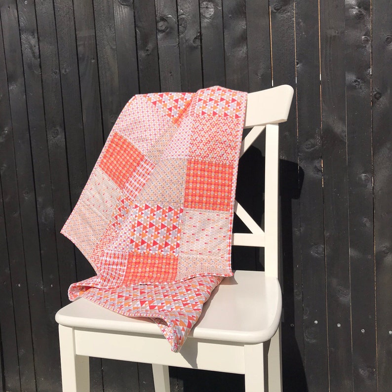 May include: A patchwork quilt in shades of pink, orange, and white, draped over a white chair. The quilt has a geometric pattern and is made of different fabrics.