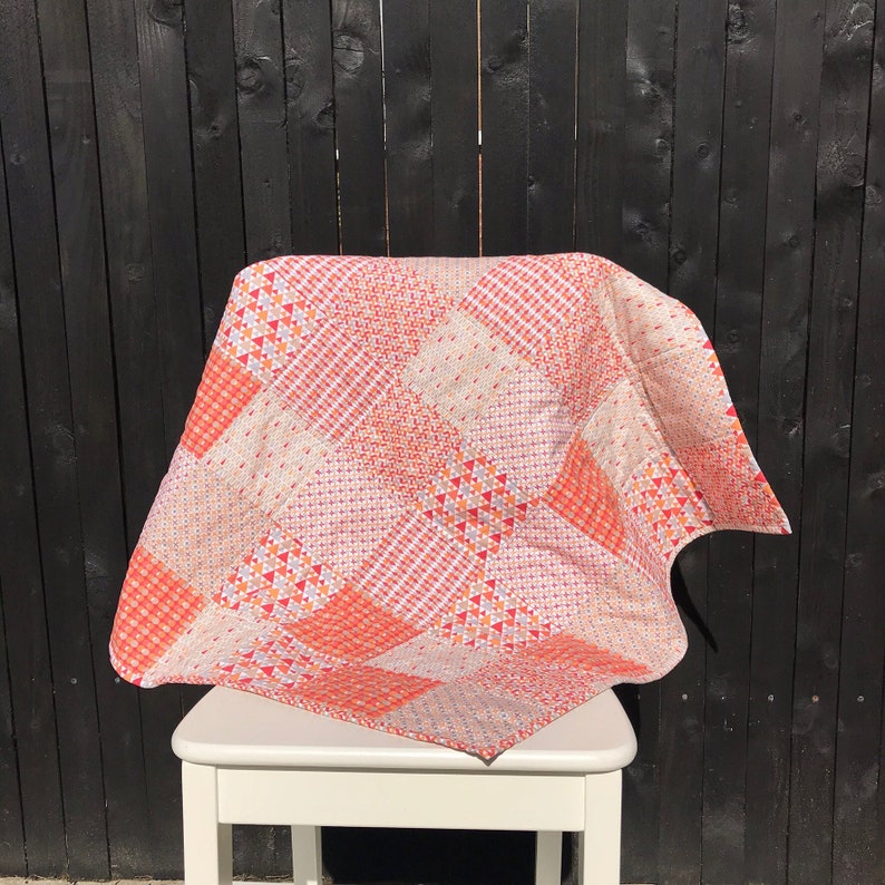 May include: A patchwork quilt with a geometric pattern in shades of orange, pink, and white. The quilt is folded and draped over a white chair.