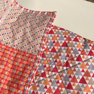 May include: A patchwork quilt with a variety of geometric patterns in shades of orange, red, gray, and beige. The quilt is made with different fabrics and has a stitched border.