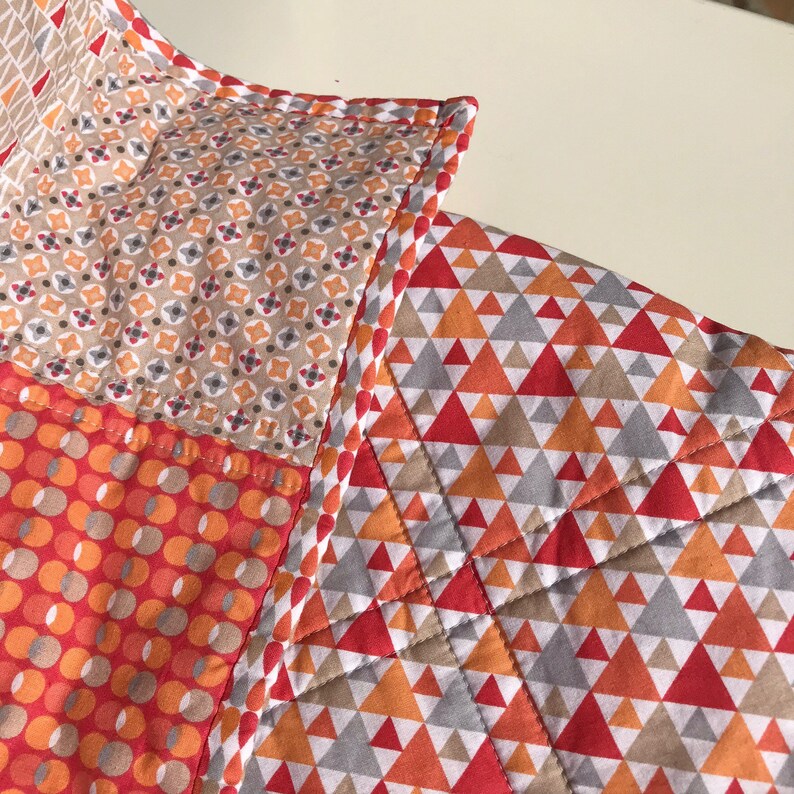 May include: A patchwork quilt with a variety of geometric patterns in shades of orange, red, gray, and beige. The quilt is made with different fabrics and has a stitched border.