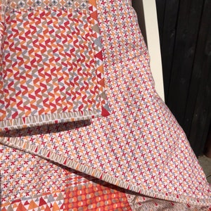 May include: A patchwork quilt with a geometric pattern in shades of red, orange, and gray. The quilt is folded and draped over a chair.