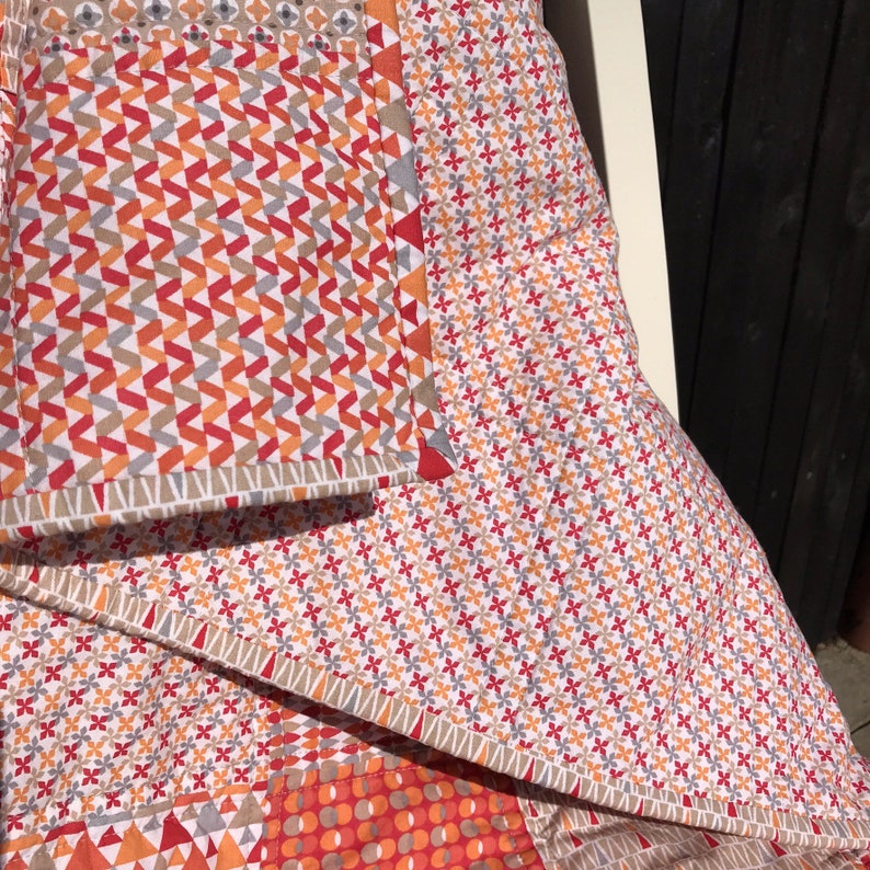 May include: A patchwork quilt with a geometric pattern in shades of red, orange, and gray. The quilt is folded and draped over a chair.