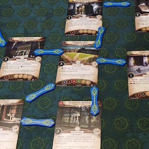 May include: A board game in play, with illustrated cards linked by blue tokens on a green patterned mat. The cards show different scenes, forming a strategic arrangement for gameplay.