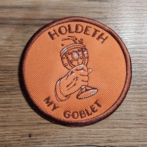 May include: Round orange patch with brown trim featuring the text "HOLDETH MY GOBLET" in a serif font. The patch depicts a hand holding a decorative goblet. The patch is likely an embroidered item.