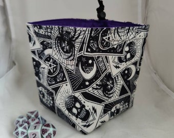 May include: A dice bag with a black and white occult print, featuring skulls and spiders. It has a drawstring closure and a purple lining. A set of dice is visible beside the bag. The bag's measurements are in centimetres.