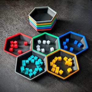 May include: A stack of six multi-colored, hexagonal trays with black interiors. Five trays are arranged around the stack, each holding small, colored cubes. The cubes are red, white, blue, teal, and yellow. The trays are on a dark wooden surface.
