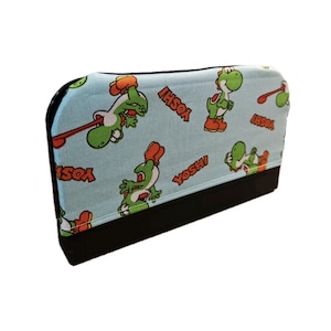 Könnte beinhalten: Eine hellblaue Hülle mit einem sich wiederholenden Muster der Figur Yoshi aus der Super Mario-Videospielserie. Das Design umfasst grüne Yoshis mit roten Schuhen und das Wort "Yoshi" in Rot. Der Boden ist schwarz.