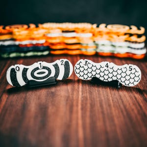 Può includere: Due segnalini di punteggio in plastica bianca con numeri e motivi neri. Il primo segnalibro ha un motivo a bersaglio e i numeri 0, 1 e 2. Il secondo segnalibro ha un motivo a scala e i numeri 3, 4 e 5.