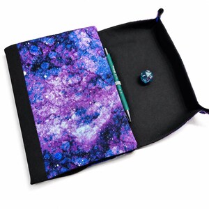 May include: A foldable fabric dice tray featuring a vibrant purple and blue galaxy print on the exterior and a solid black interior. The tray includes a teal pen with the text +1 of Writing and a single twenty-sided die resting inside.