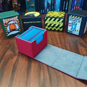 May include: A red deck box with a light grey interior containing a stack of blue cards. In the background, four other deck boxes are visible: a green one with a goose and the text PATHETIC, a black one with a wolf, a yellow and black striped one with the text WARNING! Contains Bad Decisions, and a black one with the text COUNTERSPELL CONTAINMENT UNIT.