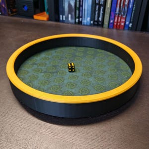 May include: A black and yellow dice tray with a green patterned interior. The tray has a black base and a yellow rim. A black die with yellow dots rests in the center of the tray. The tray is designed for tabletop gaming.