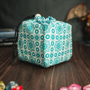 Op de afbeelding: Een vierkante dobbelsteenzak met een wit en turquoise geometrisch patroon. De stoffen tas heeft een zwarte trekkoordsluiting. Hij ligt op een donker houten oppervlak, met kleurrijke dobbelstenen en spel fiches op de voorgrond. Het patroon van de tas heeft bloemvormen.
