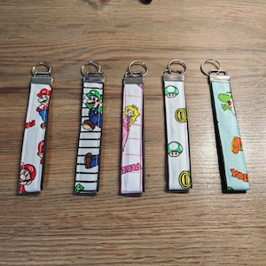 May include: Five fabric wristlet keychains featuring Nintendo character prints. The first shows Mario, the second Luigi on stripes, the third Princess Peach on a grid, the fourth green mushrooms and gold coins, and the fifth Yoshi on light blue.