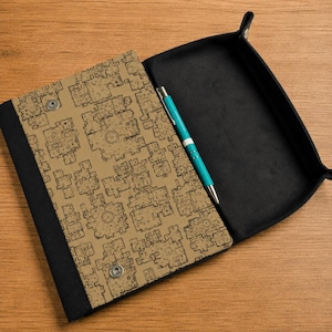 May include: A rectangular folding tray with a tan exterior featuring a black architectural floor plan pattern and a solid black interior. The tray has silver metal snap buttons at the corners and contains a single turquoise ballpoint pen.