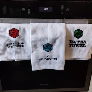 以下が含まれることがあります： 刺繍されたサイコロとテキストが施された白いキッチンクロス3枚。最初のクロスには、赤い20面ダイスで「Roll for Wetness」と書いてあります。2番目のクロスには、青い20面ダイスで「+1 of Drying」と書いてあります。3番目のクロスには、緑色の20面ダイスで「D&Tea Towel」と書いてあります。