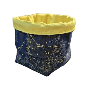 Può includere: Un cestino portaoggetti in tessuto con esterno blu navy con un motivo a costellazioni in bianco e oro. L'interno e il bordo ripiegato sono giallo brillante. Il cestino è progettato per stare in posizione verticale.